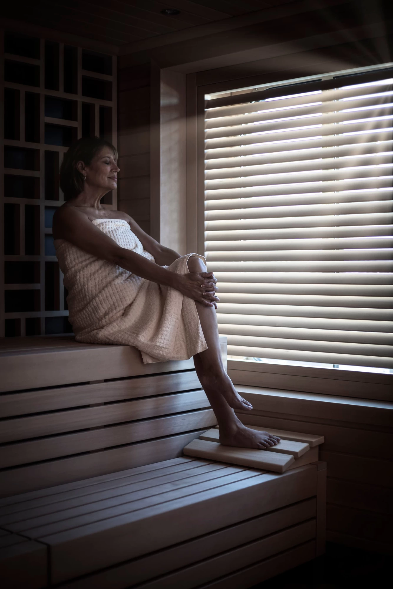 Saga cruise spa photography — woman relaxing on a wooden bench inside a sauna, side light through slatted blinds