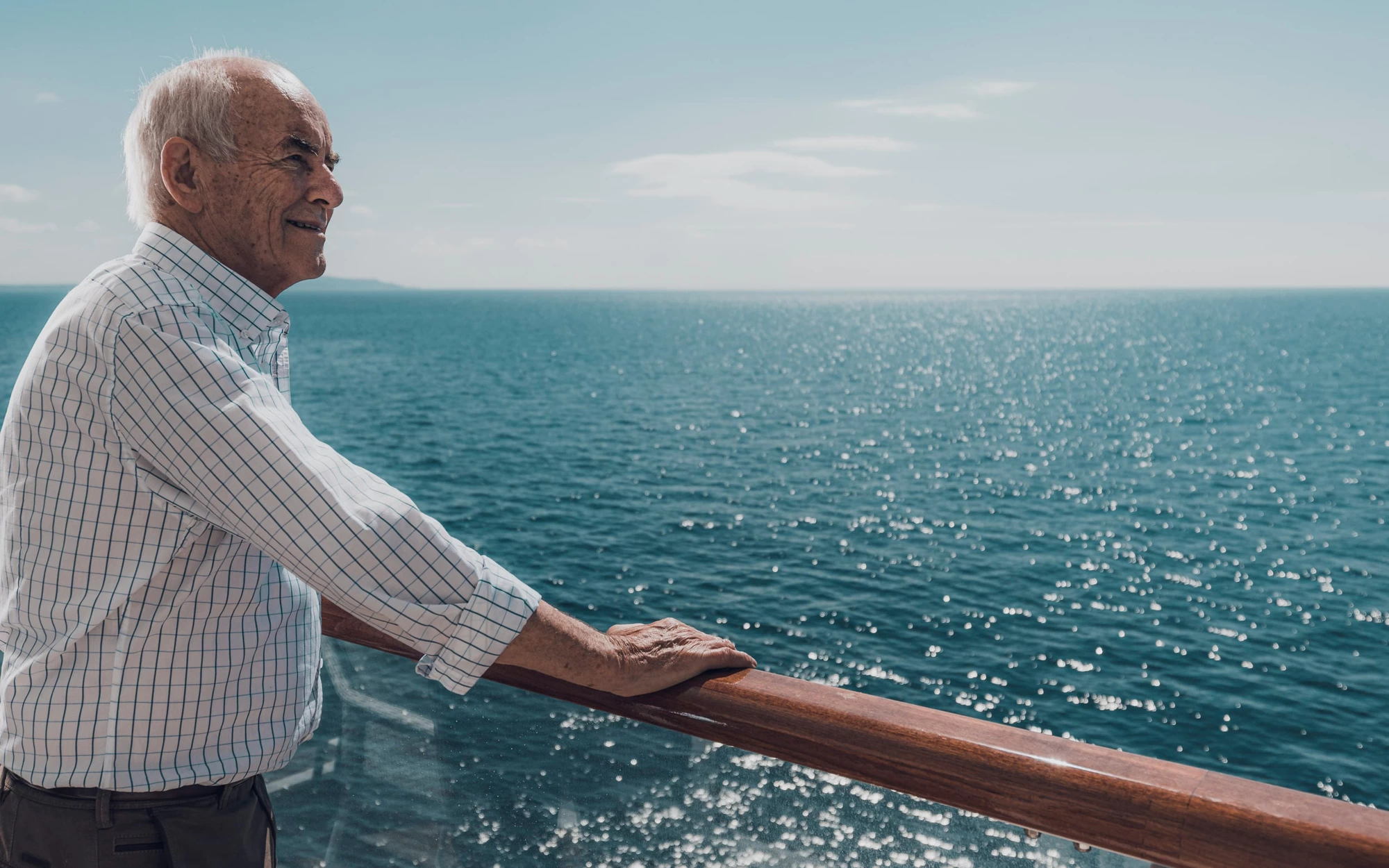 Saga cruise lifestyle photography — older man on the open deck leaning against a polished wooden rail, looking out to sea